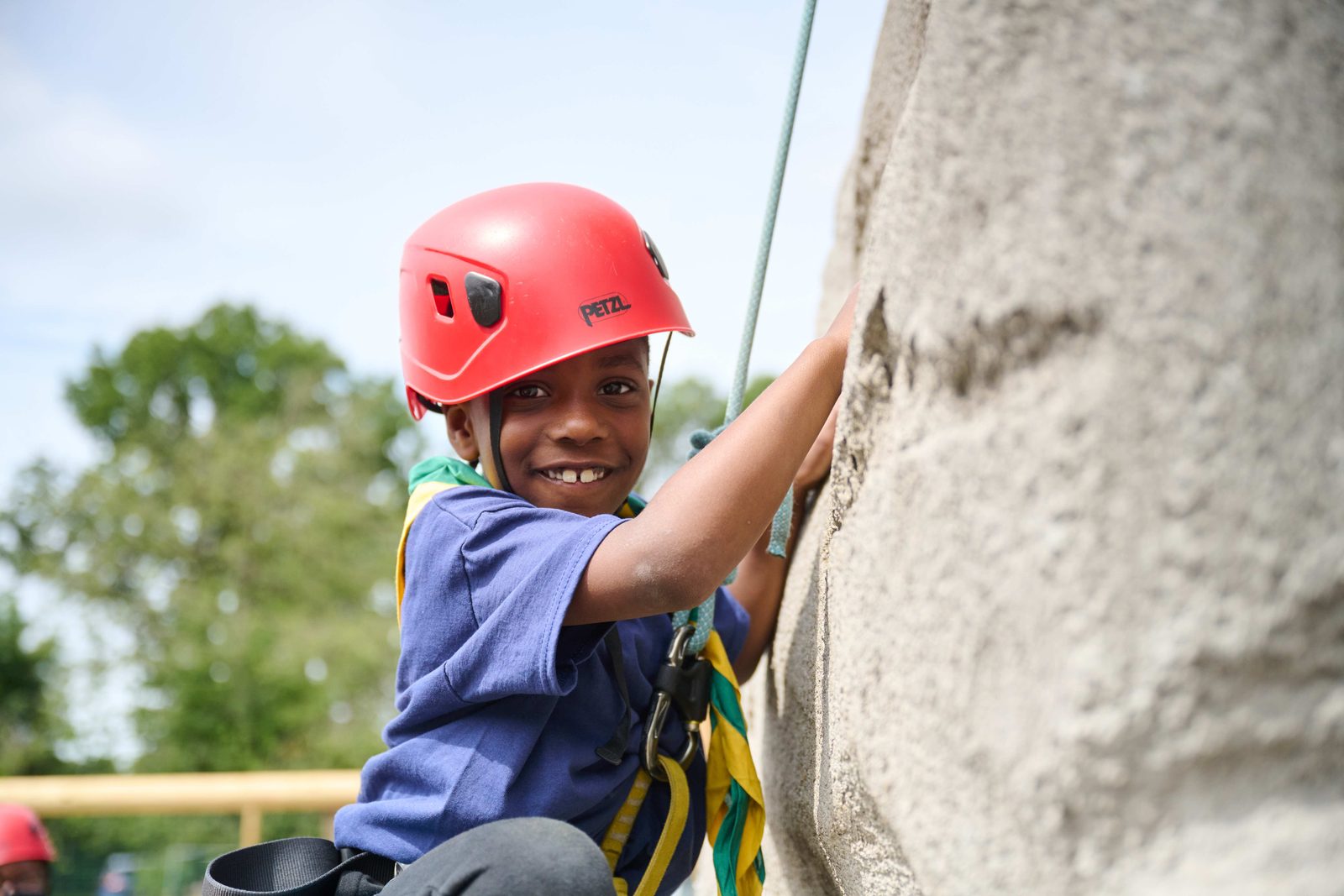 A young Scout wearing a red helmet, smiling while climbing a rock wall