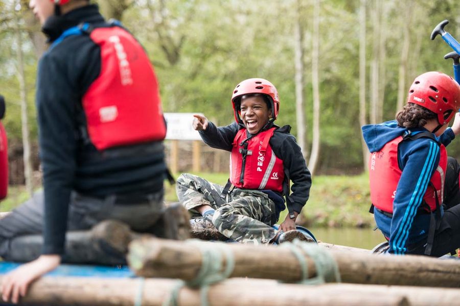 A Cub Scout laughing and pointing while rafting on a lake, wearing a helmet and life jacket