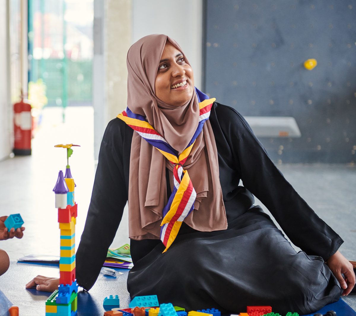 A volunteer in a Scout neckerchief sitting on the floor with young children, playing with colourful building blocks