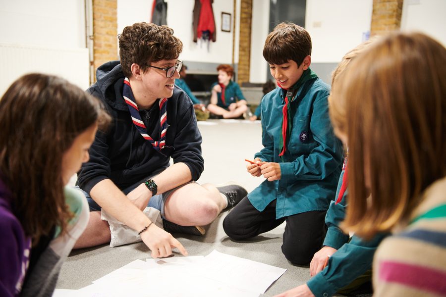 A Scout in uniform kneeling with friends, sharing programme ideas with a leader