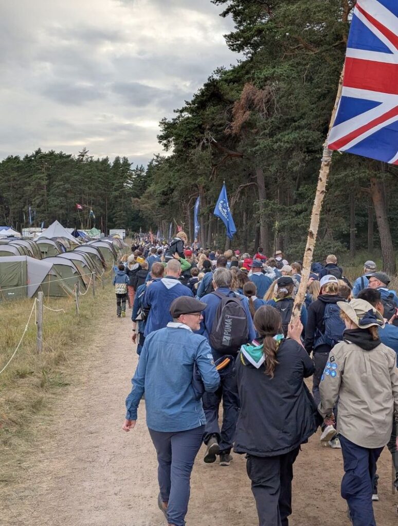 Scouts and leaders walking through camp at Jamboree26, carrying a Union Flag